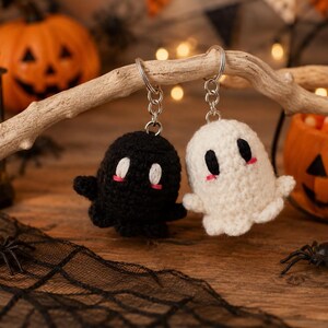 May include: Two crochet ghost keychains, one black and one white, each featuring oval eyes and small pink stitched cheeks. The figures are attached to silver-toned metal key rings, showcasing a soft, textured yarn construction with rounded shapes.