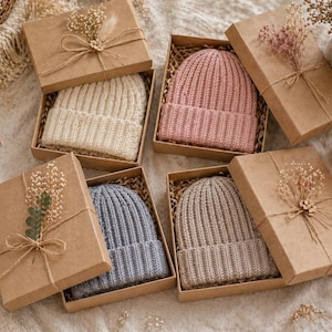 May include: Several knit beanies in various colors, including cream, pink, blue, and taupe, are presented in individual brown gift boxes. Each box is adorned with dried flowers and tied with twine, creating a rustic and elegant presentation.