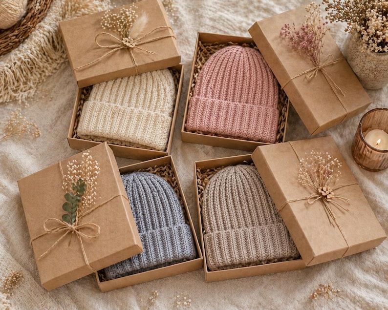 May include: Several knit beanies in various colors, including cream, pink, blue, and taupe, are presented in individual brown gift boxes. Each box is adorned with dried flowers and tied with twine, creating a rustic and elegant presentation.