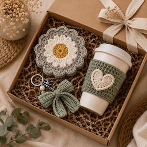 May include: A gift box containing a grey and white crocheted daisy coaster, a sage green crocheted cup sleeve with a cream heart appliqué on a white cup, and a sage green crocheted bow keyring with a silver metal ring and small beads.