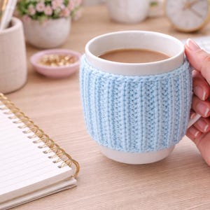 May include: A white ceramic mug filled with a light brown beverage, wrapped in a light blue knitted cozy. The mug is held by a hand. A gold pen and spiral notebook are on the wooden surface.