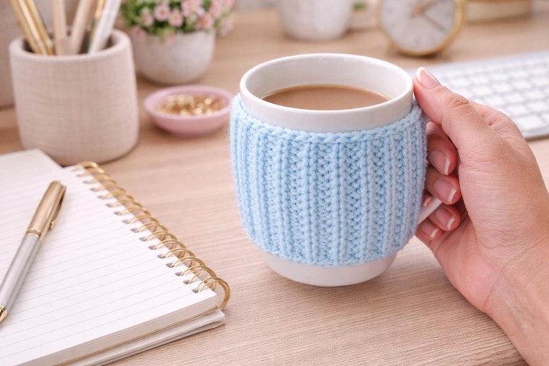 May include: A white ceramic mug filled with a light brown beverage, wrapped in a light blue knitted cozy. The mug is held by a hand. A gold pen and spiral notebook are on the wooden surface.