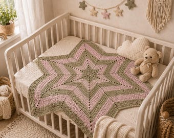 May include: A handmade star-shaped baby blanket in a crib. The blanket features alternating stripes of olive green and light pink. A beige teddy bear and cloud-shaped pillow are on the crib. The crib is white and has a white mattress.