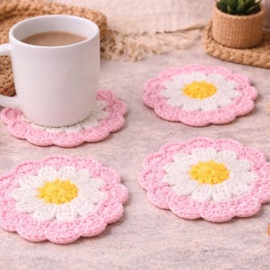 May include: A white ceramic mug filled with a beverage rests on a pink, white, and yellow crocheted daisy coaster. Three additional daisy coasters are arranged nearby. The coasters are approximately 4 inches in diameter.