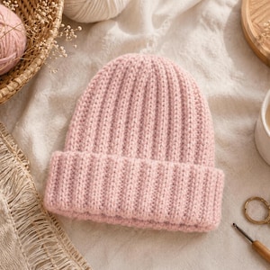May include: A light pink, ribbed knit beanie hat with a folded brim. The hat is made of a soft, textured yarn. The image also includes a basket of pink yarn, a cup of coffee, and crafting tools.