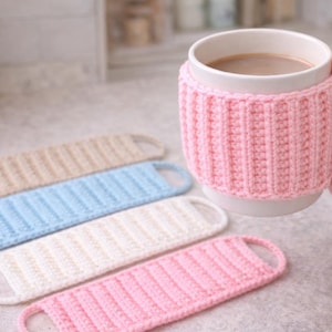 May include: A white mug with a pink knitted cozy and several matching coasters in beige, blue, white, and pink. The mug contains a warm beverage. The coasters have a loop handle.