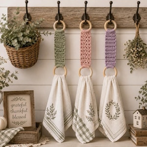 May include: Three decorative hand towels with wooden ring holders, in green, pink, and purple. Each towel is cream-colored with a green leaf design and hanging from a wooden rack. A basket of greenery and a framed sign are also visible.