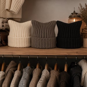 May include: Three ribbed knit beanies with pointed cat-ear silhouettes, arranged in a row on a wooden shelf. The beanies are colored in cream, taupe, and black, each featuring a wide folded cuff and a vertical knit pattern throughout the fabric.