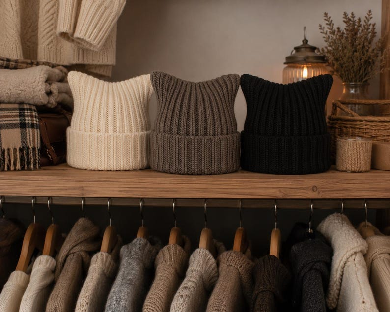 May include: Three ribbed knit beanies with pointed cat-ear silhouettes, arranged in a row on a wooden shelf. The beanies are colored in cream, taupe, and black, each featuring a wide folded cuff and a vertical knit pattern throughout the fabric.