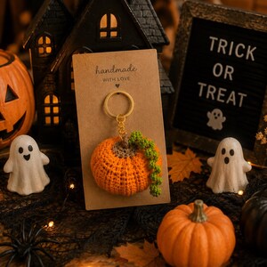 Puede incluir: Un llavero de calabaza de ganchillo naranja con una vid verde, presentado en una tarjeta marr&oacute;n que dice "handmade with love". La imagen tiene un tema de Halloween, con una casa negra, un letrero de "Trick or Treat" y otras decoraciones de calabazas y fantasmas.