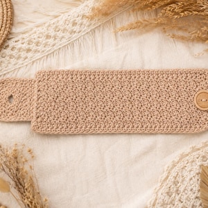 May include: A rectangular beige crochet wristband featuring a textured stitch pattern. The accessory includes a rounded tab on one end with a buttonhole and a single circular wooden button attached to the opposite end for secure fastening.