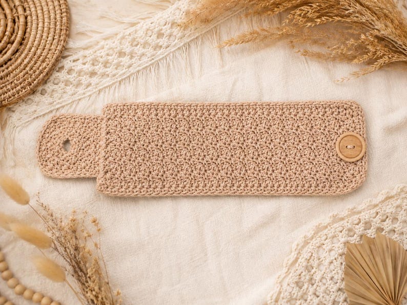 May include: A rectangular beige crochet wristband featuring a textured stitch pattern. The accessory includes a rounded tab on one end with a buttonhole and a single circular wooden button attached to the opposite end for secure fastening.