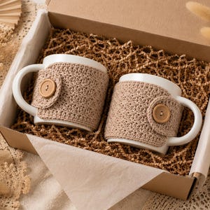 May include: Two white ceramic mugs nestled in a cardboard gift box filled with brown paper crinkle shred. Each mug features a removable beige crochet cozy secured with a single light brown wooden button on the side, providing a textured cover.
