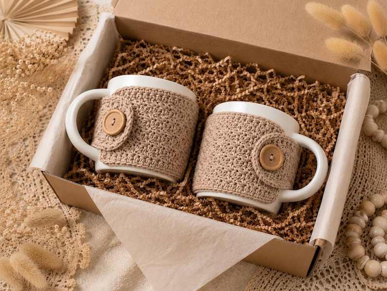 May include: Two white ceramic mugs nestled in a cardboard gift box filled with brown paper crinkle shred. Each mug features a removable beige crochet cozy secured with a single light brown wooden button on the side, providing a textured cover.