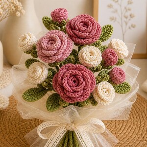 May include: A handcrafted bouquet of crochet flowers, featuring pink and cream roses with green leaves, wrapped in white tulle and tied with a cream ribbon. The bouquet is presented on a woven mat, suggesting a decorative or gift item.