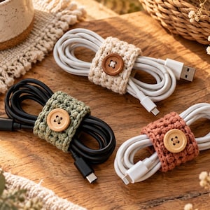 May include: Three coiled charging cables secured with crocheted cord organizers. One white cable has a beige wrap with a brown button, one black cable has a sage green wrap with a light wood button, and one white cable has a terracotta wrap with a light wood button.