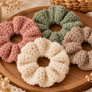 May include: A set of four circular hair scrunchies made of crocheted yarn in muted colors including dusty pink, sage green, cream, and light brown. Each scrunchie features a textured, ribbed stitch pattern and a gathered, elasticized design.