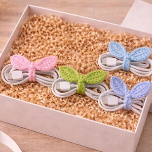 May include: A white gift box filled with shredded paper holds four white charging cables. Each cable is wrapped with a crocheted accent in pink, green, and two shades of blue, shaped like bunny ears. A ribbon and the box lid are visible.