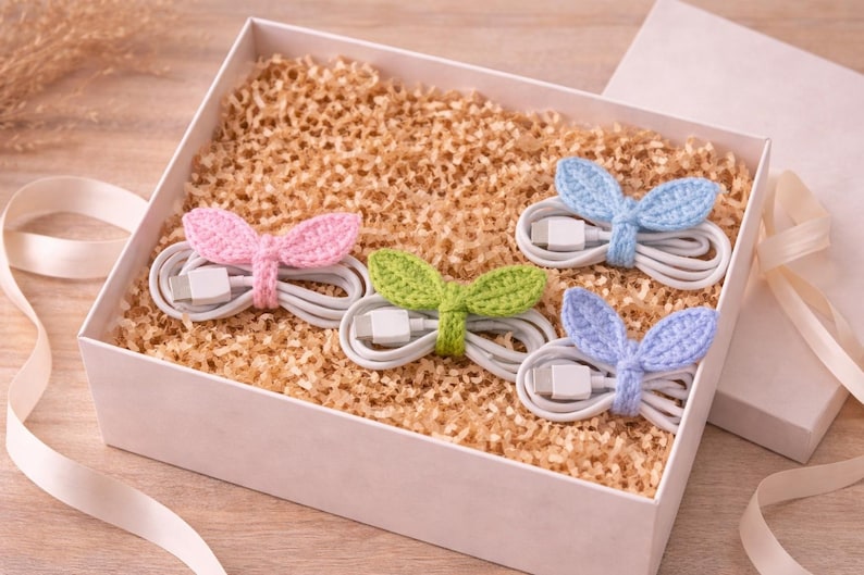 May include: A white gift box filled with shredded paper holds four white charging cables. Each cable is wrapped with a crocheted accent in pink, green, and two shades of blue, shaped like bunny ears. A ribbon and the box lid are visible.
