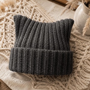 May include: A dark charcoal gray knit beanie featuring a ribbed texture and two distinct pointed corners at the top, creating a cat-ear silhouette. The hat has a wide, folded cuff at the base, constructed from a thick, soft yarn material.