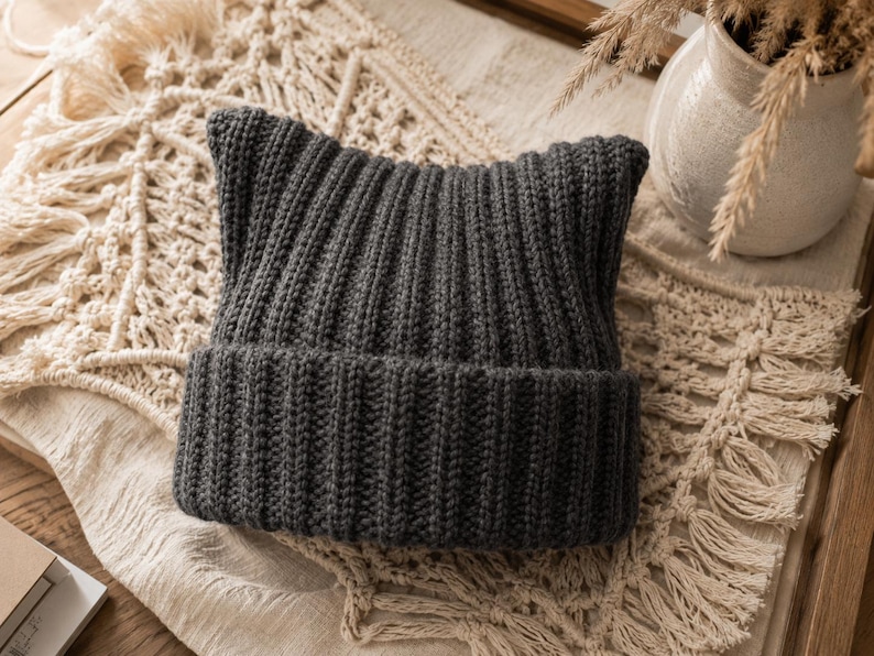 May include: A dark charcoal gray knit beanie featuring a ribbed texture and two distinct pointed corners at the top, creating a cat-ear silhouette. The hat has a wide, folded cuff at the base, constructed from a thick, soft yarn material.