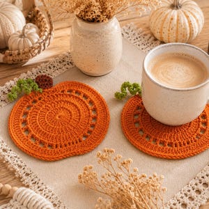May include: An autumn-themed still life featuring a cup of coffee on an orange crochet pumpkin coaster. The scene includes pumpkins, a candle, and decorative elements on a wooden surface, creating a cozy fall ambiance.