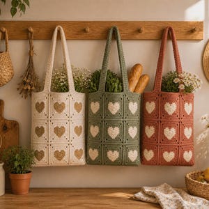Op de afbeelding: Drie gehaakte draagtassen in crème, olijfgroen en terracotta, elk met hartvormige patronen. De tassen hangen aan een houten rek, gevuld met groen en brood. Een strohoed en andere keukenartikelen zijn ook zichtbaar.