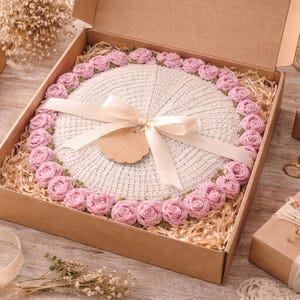 May include: A handmade, round, off-white crocheted item with a pink rose border, presented in a brown box with decorative straw. The item is tied with a cream ribbon and a small tag. Several wrapped gifts and a lit candle are also present.
