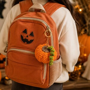 Puede incluir: Mochila naranja con un aplique de cara de calabaza. Un llavero de calabaza de ganchillo cuelga de la cremallera. La mochila tiene un bolsillo frontal y un bolsillo lateral de malla. Tema de Halloween.