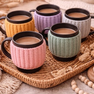May include: A set of three ceramic mugs with a speckled, cream-colored glaze. Each mug features a different hand-painted floral design in shades of blue, green, and yellow. The mugs have a simple, rounded shape and a comfortable handle.