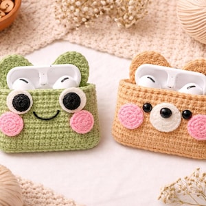 May include: Two crochet AirPod cases, one green frog-shaped and one tan bear-shaped, each holding white AirPods. The frog case has black and white eyes and pink cheeks. The bear case has black eyes and a white nose with pink cheeks. Both have ear-shaped tops.