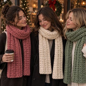 May include: Three long, chunky knit scarves in solid colors of dusty rose, cream, and sage green. Each scarf features a textured, repeating vertical stitch pattern with a soft, thick appearance, draped loosely around the neck over winter coats.