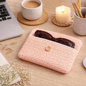 May include: A light pink crocheted glasses case with a button closure, holding a pair of tortoiseshell glasses. The case sits on a wooden desk next to a laptop, coffee, and a lit candle.