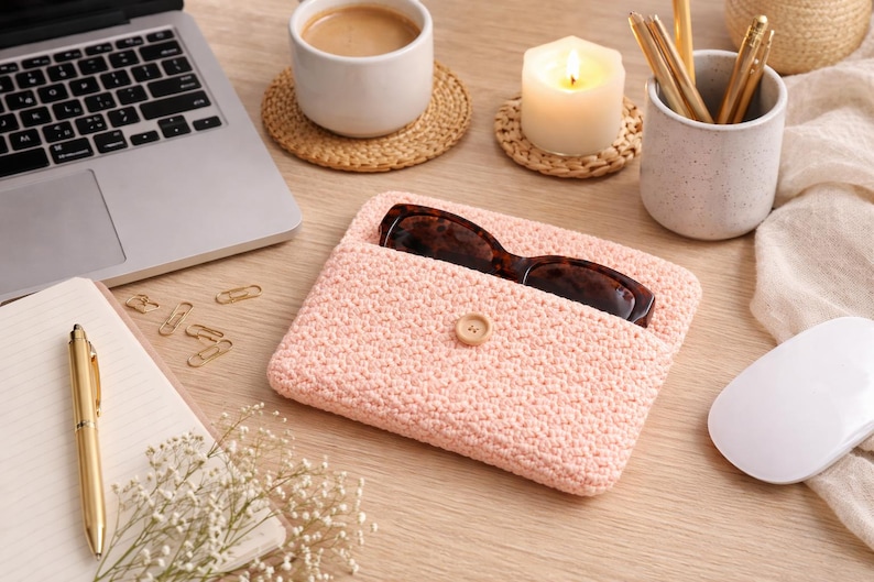 May include: A light pink crocheted glasses case with a button closure, holding a pair of tortoiseshell glasses. The case sits on a wooden desk next to a laptop, coffee, and a lit candle.
