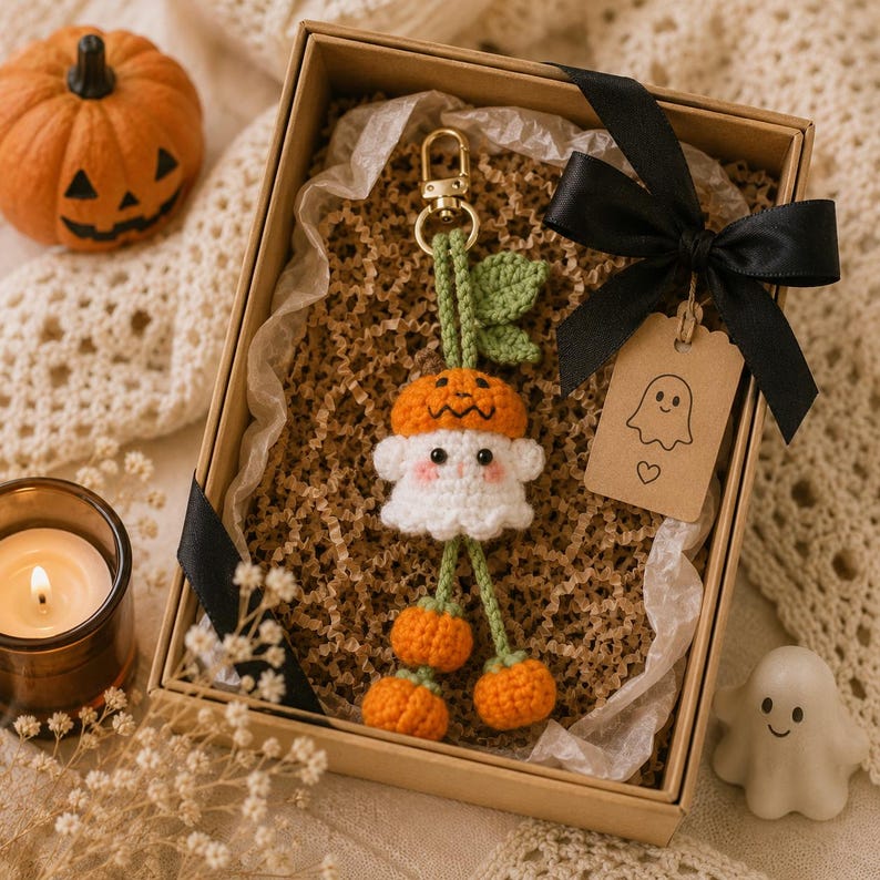 May include: A handmade Halloween-themed keychain featuring a crocheted ghost wearing an orange pumpkin hat. The keychain has green stems and orange pumpkin feet. It is presented in a gift box with a black ribbon and a tag with a ghost illustration.