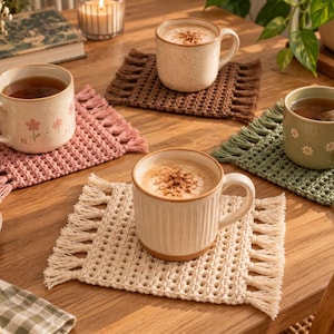 May include: Four rectangular crochet coasters with fringe edges in cream, dusty pink, sage green, and dark brown. Each coaster supports a ceramic mug filled with a beverage, featuring various textures and patterns including floral designs and ribbed sides.