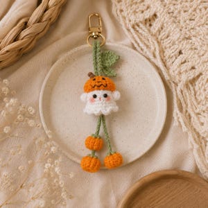 May include: A handmade Halloween-themed keychain featuring a crocheted ghost wearing a pumpkin hat. The ghost has rosy cheeks and is attached to a gold-colored clasp. Two orange crocheted pumpkins dangle below the ghost. The keychain is approximately 4 inches long.