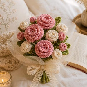 May include: A handcrafted bouquet of crochet flowers, featuring pink and cream-colored roses with green leaves. The bouquet is wrapped in a sheer, cream-colored fabric and tied with a matching bow. The flowers are arranged to resemble a natural bouquet.