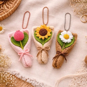 May include: Three handmade crochet flower bouquets: a pink tulip, a yellow sunflower, and a white daisy. Each bouquet is wrapped in a matching color and tied with a ribbon. They have a small chain for hanging.