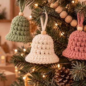 May include: Three crochet bell ornaments hanging from a Christmas tree. The bells are sage green, cream, and dusty rose, each featuring a textured stitch pattern, a small wooden bead at the top, and a matching yarn loop for hanging on a branch.
