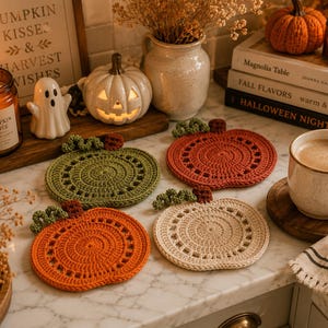 May include: A cozy autumn-themed display featuring four crocheted pumpkin coasters in orange, green, and cream. The scene includes lit candles, a pumpkin-shaped light, and a sign that reads "PUMPKIN KISSES & HARVEST WISHES."