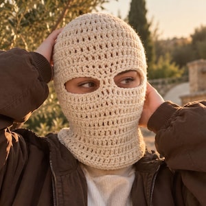 May include: A cream-colored crochet balaclava featuring a textured, open-stitch pattern with two distinct eye openings. The headgear is crafted from yarn and covers the head and neck, providing a snug fit around the face and forehead.