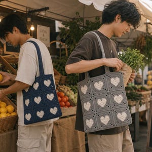 May include: Two crochet tote bags with heart patterns. One bag is navy blue with white hearts, the other is gray with white hearts. The bags have square panels and long straps. The bags are being carried at a farmer's market.