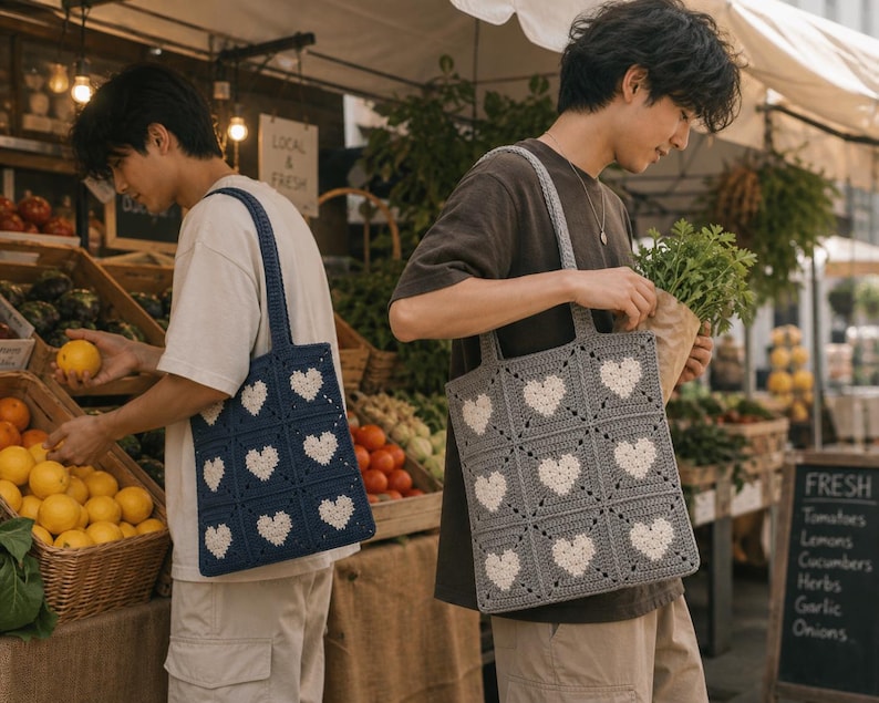 May include: Two crochet tote bags with heart patterns. One bag is navy blue with white hearts, the other is gray with white hearts. The bags have square panels and long straps. The bags are being carried at a farmer's market.