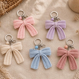 May include: A collection of six handmade crochet bow keychains in various pastel colors: pink, lavender, cream, blue, and coral. Each bow is attached to a silver keyring with a decorative bead. The keychains are arranged on a neutral-toned surface.