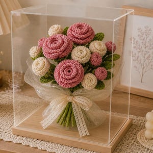May include: A handcrafted crochet bouquet of pink and cream-colored roses and buds, with green leaves, presented in a clear display case. The bouquet is tied with a cream-colored ribbon. The display case sits on a wooden base.