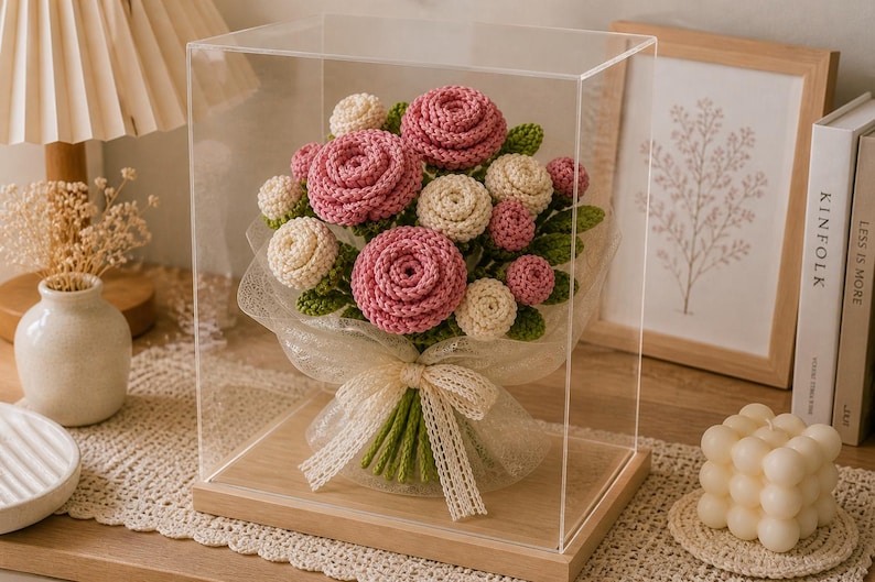 May include: A handcrafted crochet bouquet of pink and cream-colored roses and buds, with green leaves, presented in a clear display case. The bouquet is tied with a cream-colored ribbon. The display case sits on a wooden base.