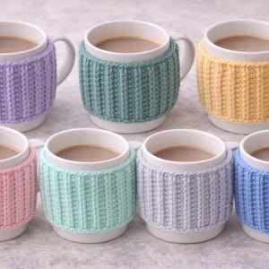 May include: Seven white ceramic mugs filled with a warm beverage, each adorned with a knitted cozy. The cozies come in various pastel colors: lavender, teal, yellow, pink, mint green, gray, and blue. The mugs have white handles.