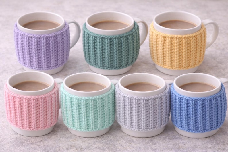 May include: Seven white ceramic mugs filled with a warm beverage, each adorned with a knitted cozy. The cozies come in various pastel colors: lavender, teal, yellow, pink, mint green, gray, and blue. The mugs have white handles.