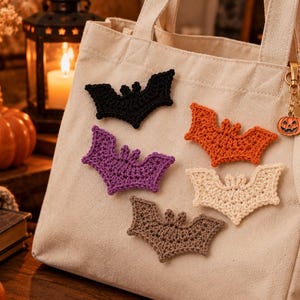 May include: A beige canvas tote bag decorated with five crocheted bat appliques in black, purple, orange, cream, and brown. A small gold-toned metal keychain featuring a carved pumpkin charm hangs from the side of the bag handle.